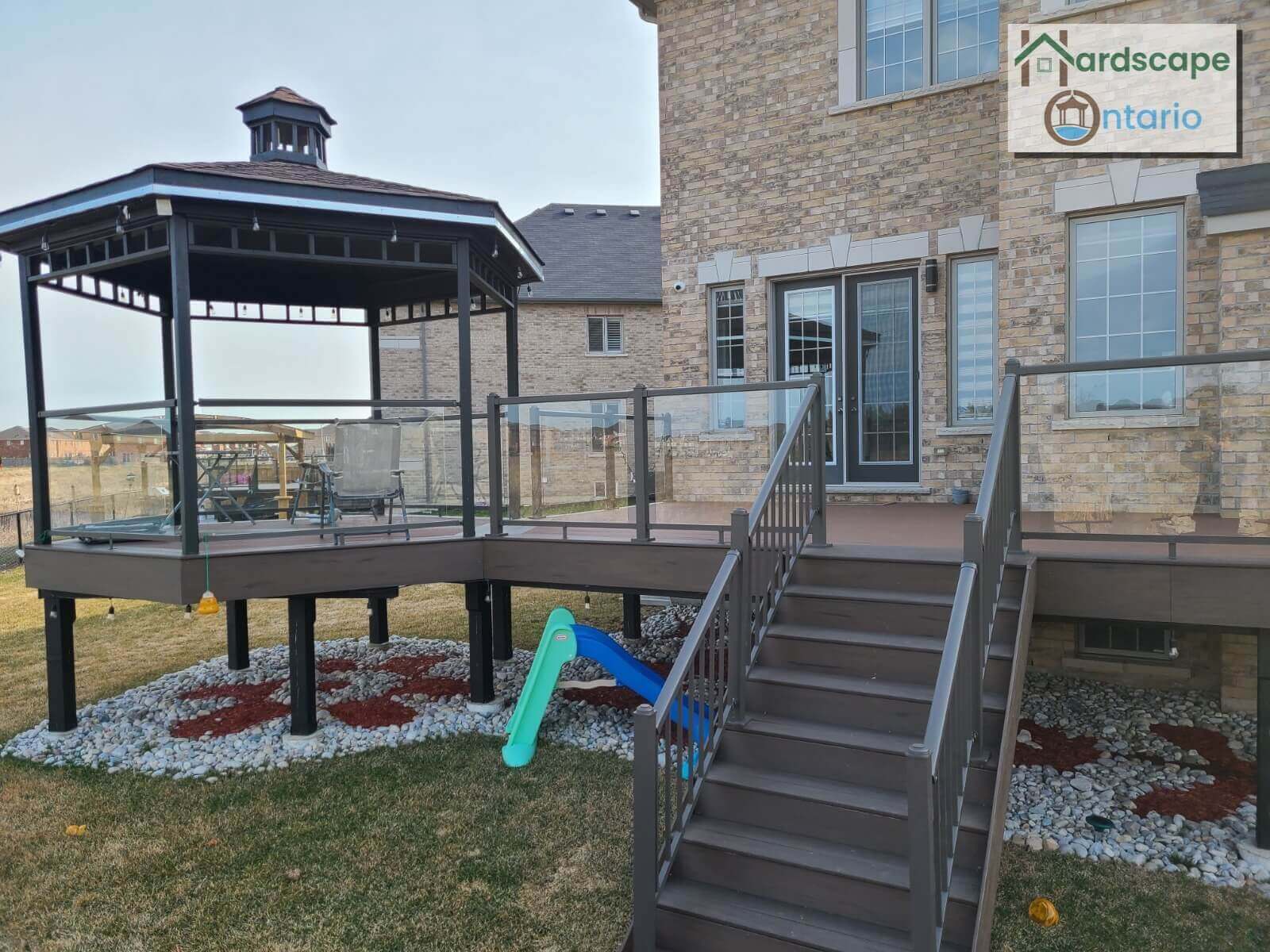 Hexagonal shaped gazebo with an incredible designed roof and tempered glass bars for protection also levelled wooden deck with tinted glass bar screens for protection with some wooden stairs. Under the deck is designed with river rock and brown mulch.